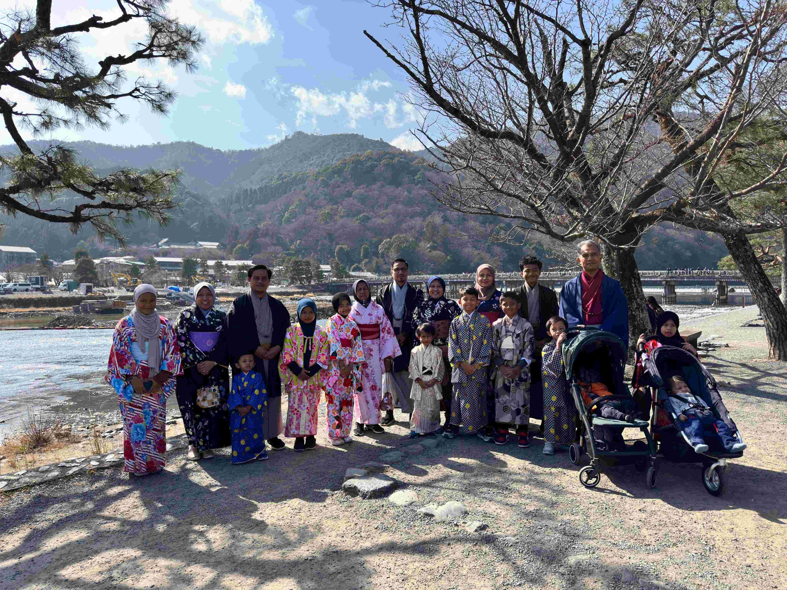 A large family group, including women wearing hijabs, dressed in colorful traditional Japanese kimonos and yukatas, posing together by the river in Arashiyama, Kyoto, with mountains in the background.